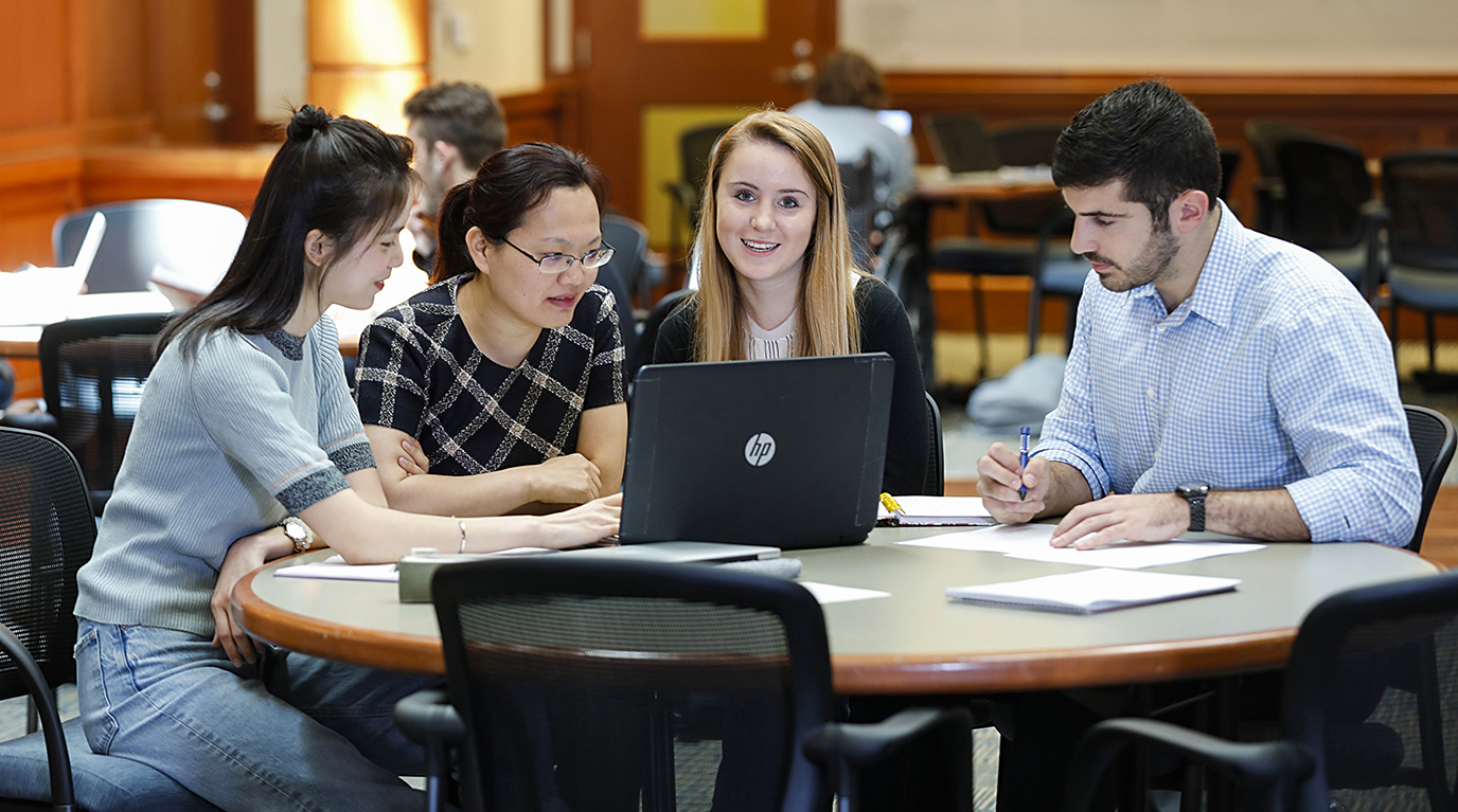 4 students studying at a table in one of the college's study spaces. There are 3 people looking down at notes and their laptop and the fourth, in the center of the photo, is smiling at the camera.